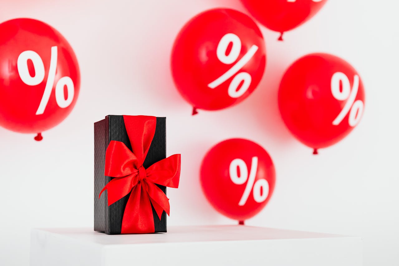 A Gift With Red Ribbon in Between Red Balloons With Percentage Symbols on a White Background